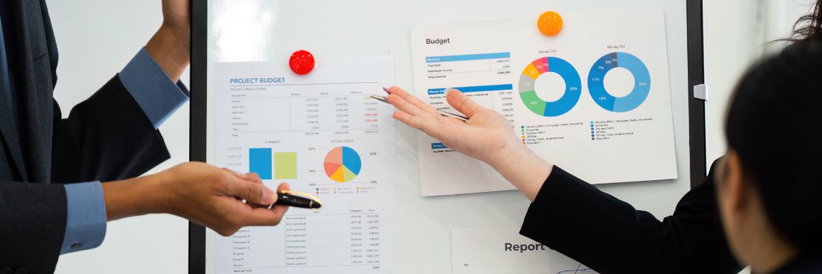 Two people pointing at printed budget reports and pie charts pinned to a whiteboard during a presentation.
