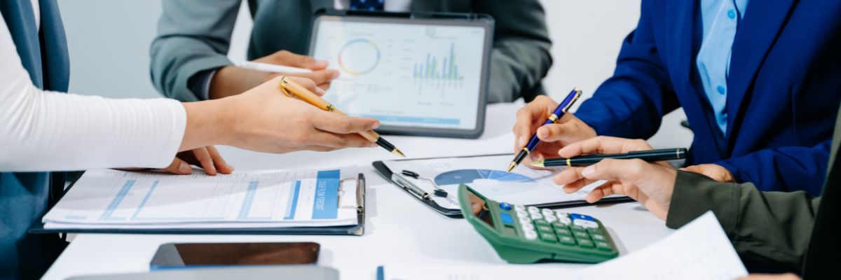 Hands pointing at various financial charts and a calculator on a desk, with a tablet displaying more data in the background.