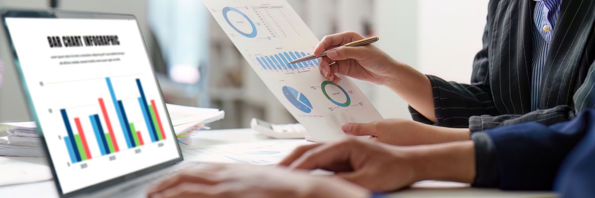 Hands reviewing printed charts next to a laptop showing a bar chart infographic.