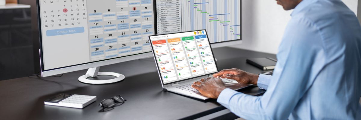 A person using a laptop to manage a digital Kanban board with multiple monitors showing project calendars and Gantt charts in the background.